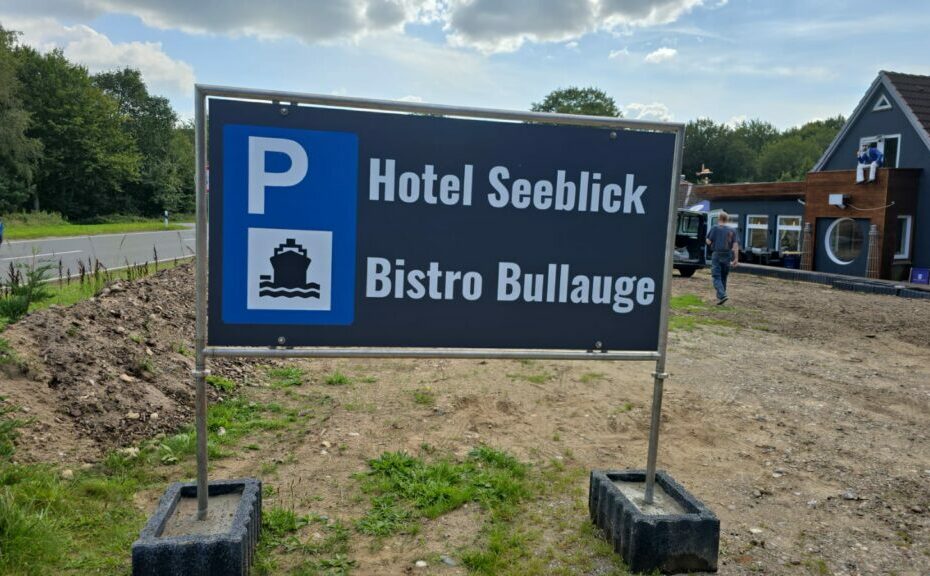 Auf einem Schild im Außenbereich sind die Informationen zum „Hotel Seeblick“ und zum „Bistro Bullauge“ mit einem Parkplatzsymbol (Hinweis: Parkplatz) zu sehen. Im Hintergrund sind eine Schotterfläche und ein Gebäudeteil zu sehen, außerdem ein blaues Auto und etwas Grün.